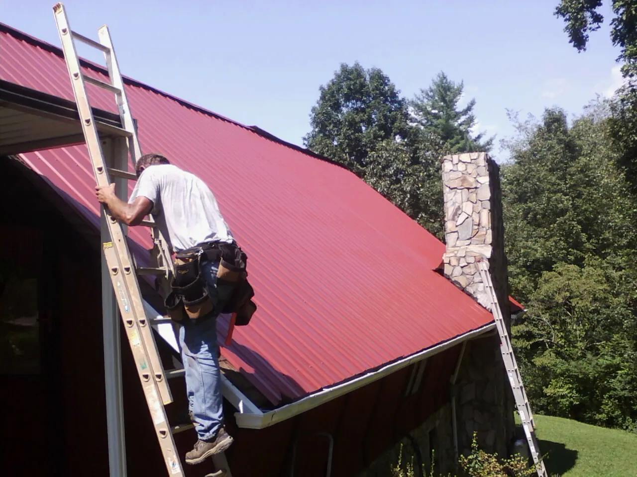Roofer on a ladder beside a red metal roof and stone chimney — Soffit & Fascia Repair by Roofing Brothers in Lawrenceburg